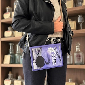 Puede incluir: Un bolso cruzado morado con el texto "Advanced Potion Making" en la parte delantera. El bolso tiene un diseño de libro con una silueta negra y una correa de color claro. La persona lleva una chaqueta de cuero negra y vaqueros oscuros.