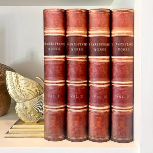 Peut inclure: Quatre volumes des Œuvres de Shakespeare sont présentés sur une étagère. Les livres ont un aspect vintage avec une couverture en cuir rouge foncé et marron. Les dos sont étiquetés avec le titre et les numéros de volume.