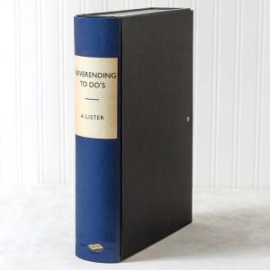May include: A black storage box with a blue spine designed to look like a book. The spine features the text "NEVERENDING TO DO'S" and "A. LISTER" on a tan label. The box is on a white surface.