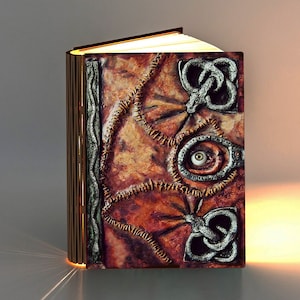 May include: A book-shaped lamp with a brown leather-like cover, intricate metal details, and a single eye peering out from the center. The lamp is illuminated from within, casting a warm glow.