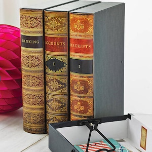 May include: Three storage boxes designed to look like antique books, labelled "BANKING", "ACCOUNTS", and "RECEIPTS". The spines are black, red, and brown with gold detailing. A fourth grey box is visible, with an open interior.
