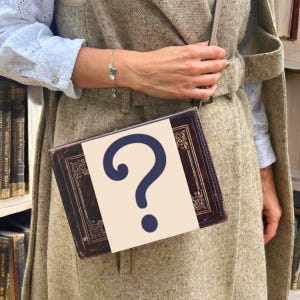 Puede incluir: Un bolso con forma de libro de cuero marrón con un gran signo de interrogación azul sobre un rectángulo crema. El bolso lo lleva una persona que lleva un abrigo beige y una blusa blanca. Se ve una pulsera plateada con detalles verdes.