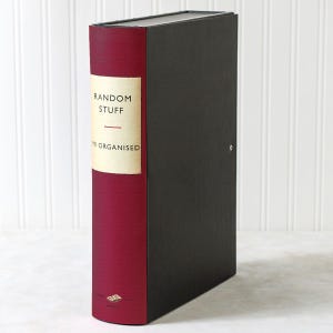 Op de afbeelding: Een zwarte opbergdoos in de vorm van een boek met een donkerrode rug. De rug is voorzien van een crèmekleurig label met de zwarte tekst "RANDOM STUFF" en "MR ORGANISED", en een klein logo onderaan de rug.