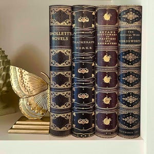 Puede incluir: Una figura de mariposa de metal dorado se encuentra junto a cuatro libros antiguos con lomos de cuero oscuro y letras doradas en relieve. Los libros se titulan Smollett's Novels, Thackeray's Works, Bryan's Dictionary of Painters and Engravers y The Poetical Works of Wordsworth.
