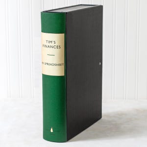 May include: A vertical green and black storage box designed to resemble a book. The green spine features a cream-coloured label with the black text "TIM'S FINANCES" and "MR SPREADSHEET" printed on it, along with a small penguin logo at the bottom.