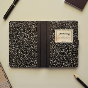 Puede incluir: Una funda rectangular para cuaderno con un patrón moteado en blanco y negro. La funda tiene un lomo central negro y una etiqueta blanca a la derecha con el texto COMPOSITIONS y el nombre H. Granger escrito a mano en la línea.