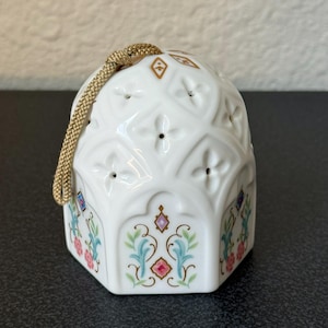 May include: A white ceramic bell ornament featuring a raised diamond pattern with small cutouts and a gold-coloured hanging cord. The base is decorated with colourful floral motifs in pink, blue, and green, accented by small diamond-shaped designs.