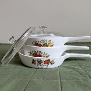 Puede incluir: Una pila de tres sartenes de cerámica blanca con mangos largos y una tapa de cristal transparente. Cada sartén presenta un estampado botánico colorido de verduras y hierbas, con el texto Le Persil impreso en negro en la sartén inferior.