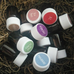 Puede incluir: Una colección de pequeñas botellas de vidrio con tapas blancas y etiquetas de colores. Las etiquetas tienen texto, incluyendo "Tea Tree", "Lavender", "Stress Away" y "Panaway".