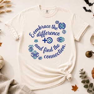May include: An ivory short-sleeve t-shirt featuring blue text that reads "Embrace the difference and find the connection." The design includes a central symbol combining a female sign with a circle and various geometric patterns in shades of blue.