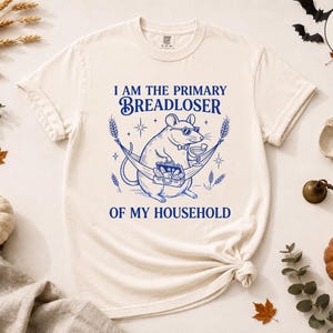 May include: An ivory t-shirt featuring a blue graphic of a rat wearing sunglasses and a bowtie, lounging in a hammock with a briefcase and a drink. The text reads: I AM THE PRIMARY BREADLOSER OF MY HOUSEHOLD. The design includes wheat stalks and stars.