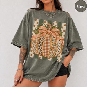 May include: A moss green oversized t-shirt featuring a large graphic of a pumpkin with a bow. The pumpkin has segments with leopard print, plaid, and floral patterns. The background of the graphic includes vertical stripes and polka dots.