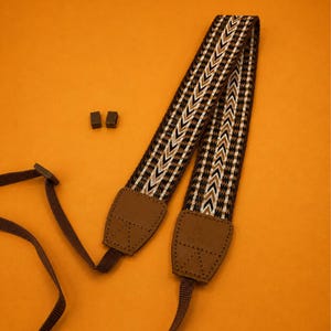 May include: A camera strap featuring a woven pattern of brown, black, and white chevron stripes. The strap includes brown leather-like end tabs with visible stitching and a brand logo, along with adjustable dark brown nylon webbing and two small black plastic sliders.