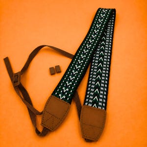 May include: A camera strap featuring a woven pattern of dark green, black, and white chevron shapes. The strap includes brown leather end tabs with stitching and brown nylon adjustment webbing, accompanied by two small brown rectangular plastic pieces.