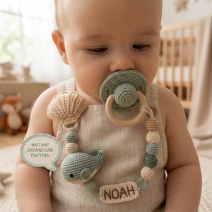 May include: A crochet pacifier clip featuring a sage green whale, a beige seashell, and wooden beads. The clip includes a rectangular tag with the name NOAH embroidered in brown thread. A speech bubble graphic states INSTANT DOWNLOAD PATTERN.