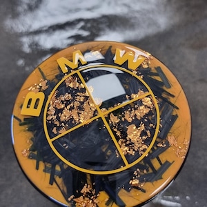 May include: A circular emblem featuring the BMW logo in yellow and black. The design includes a glossy resin finish with embedded black fiber strands and gold leaf flakes. The letters B, M, and W are raised in yellow, positioned above the central circular logo.