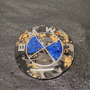 May include: A circular BMW emblem featuring a black and gray carbon fiber pattern background. The design is accented with metallic gold leaf flakes and vibrant blue foil fragments. The letters B, M, and W are displayed in a clean, white, sans-serif font.