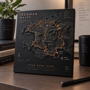 May include: A black square plaque featuring a 3D topographical map of the Ironman Wales course in Pembrokeshire. The map displays the swim, bike, and run routes with orange lines, text labels for locations, a 20 km scale, and the text YOUR NAME HERE.