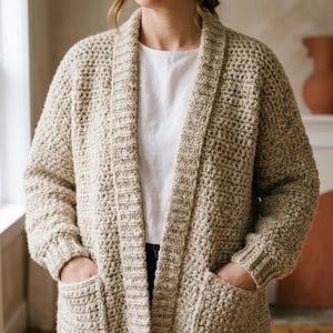 May include: A beige, chunky knit cardigan featuring a textured stitch pattern, long sleeves with ribbed cuffs, a wide ribbed collar, and two front patch pockets. The garment is worn over a plain white shirt, showcasing a relaxed, open-front design.