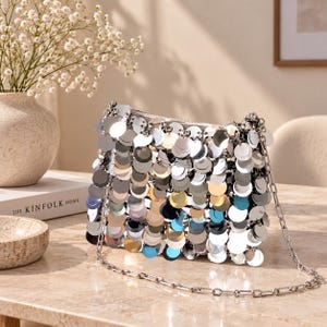 May include: A rectangular handbag covered in overlapping circular silver sequins that create a reflective, metallic surface. The bag features a thin, silver-toned metal chain strap and a secure zip closure along the top edge for functional use.
