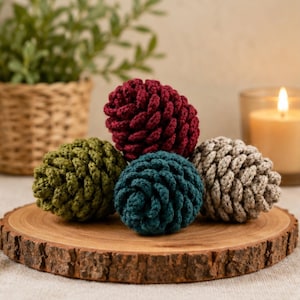 Puede incluir: Cuatro piñas de ganchillo en hilo rojo oscuro, verde oliva, azul verdoso y beige. Cada piña presenta un patrón de puntos texturizado y en capas que imita la apariencia de las escamas naturales. Los artículos están dispuestos en un grupo.