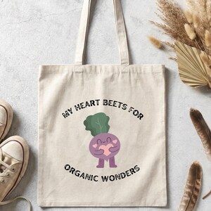 May include: A beige canvas tote bag featuring a central graphic of a purple beet character holding a pink heart. The text reads MY HEART BEETS FOR ORGANIC WONDERS in black lettering. The bag has two shoulder straps and a rectangular body shape.