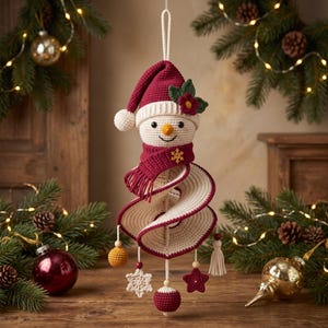 Könnte beinhalten: Ein gehäkelter Schneemann-Anhänger mit bordeauxroter Mütze und Schal sowie einem spiralförmigen Körper in Creme und Bordeaux. Die Dekoration ist mit einer kleinen goldenen Schneeflocke, einer roten Blume und hängenden Anhängern verziert.