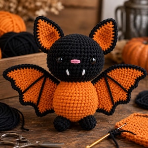 May include: A crocheted bat toy featuring a round black head, a round orange body, and large orange wings with black edges. The bat has black button eyes, a small pink stitched nose, and two white fangs protruding from the mouth area.