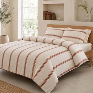 Puede incluir: Un juego de cama compuesto por una funda nórdica y dos fundas de almohada a juego, de color crema con rayas verticales de color óxido. La tela tiene un tejido texturizado visible y la funda nórdica incluye una fila de pequeños botones.