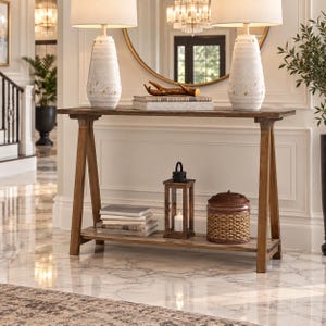 May include: A wooden console table with an A-frame base and a lower shelf. The top surface holds two white textured table lamps and a stack of books topped with a decorative antler. The lower shelf holds a wooden lantern and a woven basket.