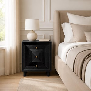 May include: A black three-drawer nightstand with a concentric square textured pattern and small gold circular knobs. A cream-colored table lamp with a cylindrical shade sits on top, next to a closed book, positioned beside a beige upholstered bed.