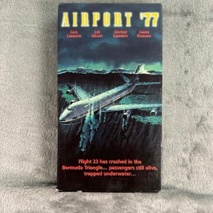 Könnte beinhalten: Ein VHS-Kassetten-Cover für den Film Airport '77 mit einem großen Passagierflugzeug, das in dunklem Ozeanwasser versunken ist. Der Titel ist oben in Gelb gedruckt, mit den Namen der Schauspieler und einer Zusammenfassung in rotem Text unten.