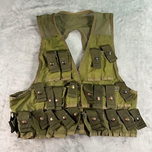 May include: An olive green tactical vest featuring a mesh upper back panel and numerous front pockets. Each pocket is secured with a flap and a metallic snap button closure. The garment is constructed with multiple rows of storage compartments.