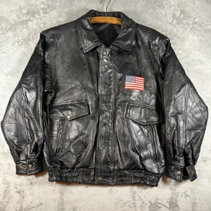 Puede incluir: Una chaqueta de cuero negra con diseño de patchwork, cuello clásico y cierre de cremallera frontal. La chaqueta incluye dos bolsillos delanteros con solapa y botones a presión, y un parche rectangular de la bandera de Estados Unidos en el pecho izquierdo.