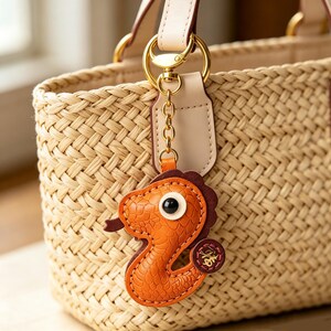 Peut inclure: Un sac cabas en paille tressée avec un porte-clés en métal doré. Le porte-clés est en forme d'hippocampe en cuir orange texturé avec une crinière marron foncé, un œil noir et blanc, et une petite étiquette ronde en cuir marron avec du texte doré.