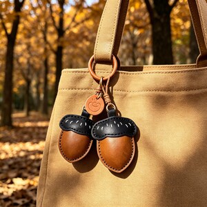 Op de afbeelding: Een beige canvas tas met twee eikelvormige lederen bedels bevestigd aan een ronde ring. Elke eikel heeft een zwarte dop met witte stiksels en een gladde bruine onderkant. Een klein rond lederen label met een lachend gezichtje is inbegrepen.