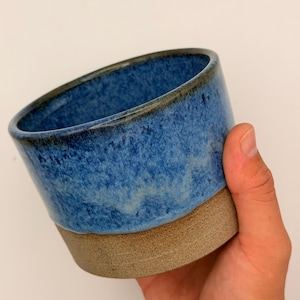 Peut inclure: Une tasse en céramique avec une partie supérieure émaillée bleu vif à la texture mouchetée et un bord foncé. La partie inférieure de la tasse est laissée dans une finition beige mate, naturelle et non émaillée, créant un contraste net.