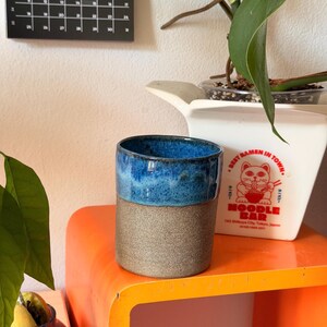 Könnte beinhalten: Ein zylindrischer Keramikbecher mit einer texturierten, matten grauen Basis und einem glänzend leuchtend blauen Rand. Der Becher hat ein schlichtes, henkelloses Design mit einem deutlichen Kontrast zwischen dem rauen unteren Teil und dem glatten oberen Teil.