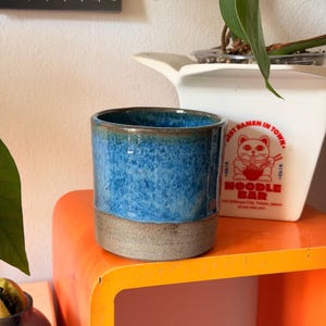 Peut inclure: Une tasse en céramique cylindrique présentant une section supérieure émaillée bleu vif avec une texture mouchetée et une base grise mate contrastante. La tasse a un design lisse à parois droites avec un bord foncé, offrant une finition bicolore.