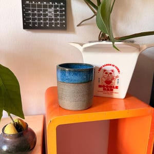 Könnte beinhalten: Ein zylindrischer Keramikbecher mit zweifarbigem Design, bestehend aus einer texturierten, gesprenkelten grauen Basis und einem glatten, leuchtend blauen glasierten Rand. Der Becher steht vor einem weißen, quadratischen Behälter mit einer roten Katzen-Grafik.