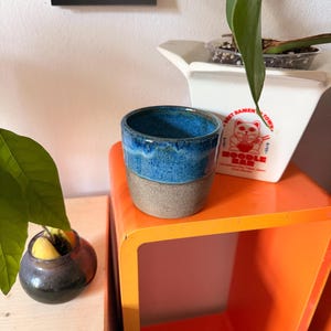 Könnte beinhalten: Ein zylindrischer Keramikbecher mit einem glänzend blau glasierten oberen Teil und einer strukturierten, matten grauen Basis. Der Becher steht auf einer orangefarbenen Oberfläche neben einem weißen Behälter mit einer roten Grafik einer Katze.