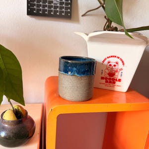 Könnte beinhalten: Ein zylindrischer Keramikbecher mit einer texturierten, hellgrau gesprenkelten Basis und einem glatten, tiefblauen glasierten Rand. Der Becher steht auf einer leuchtend orangefarbenen Oberfläche neben einem weißen Behälter mit rotem Katzenmotiv.