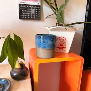 May include: A cylindrical ceramic cup with a two-tone finish, featuring a glossy blue glaze on the upper half and a matte, textured grey base. The cup sits on an orange surface next to a white container with a red graphic of a cat eating noodles.