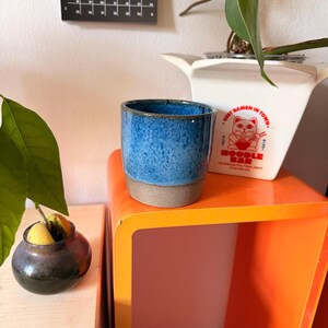 Könnte beinhalten: Ein zylindrischer Keramikbecher mit einer gesprenkelten blauen Glasur im oberen Bereich und einem matten, unglasierten grauen Boden. Der Becher steht auf einer leuchtend orangefarbenen Fläche neben einem weißen Übertopf mit rotem Katzenmotiv.