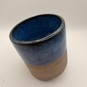 Könnte beinhalten: Ein zylindrischer Keramikbecher mit zweifarbigem Design. Der obere Teil ist mit einer glänzenden, gesprenkelten tiefblauen Glasur versehen, während der untere Teil ein rohes, strukturiertes und erdfarbenes Ton-Finish mit sichtbarer körniger Oberfläche aufweist.