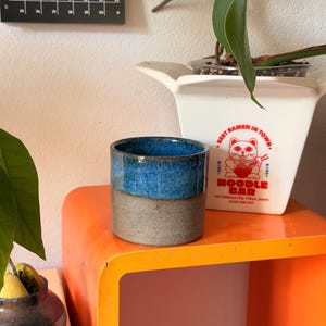 Peut inclure: Une tasse cylindrique en céramique avec un intérieur et un bord supérieur émaillés bleu profond brillant, passant à une finition grise mate texturée sur la moitié inférieure. La tasse repose sur une surface orange vif à côté d'un récipient blanc.