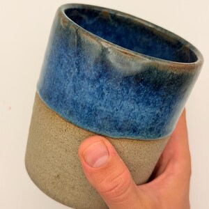 Könnte beinhalten: Ein zylindrischer Keramikbecher mit zweifarbigem Design. Der obere Teil ist mit einer glänzenden, tiefblauen Glasur versehen, die in eine strukturierte, matte beigefarbene Basis übergeht. Das Innere des Bechers ist ebenfalls tiefblau glasiert.