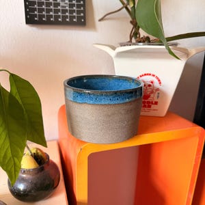 Peut inclure: Un pot de fleurs cylindrique en céramique avec une base extérieure gris mat texturée et un bord émaillé bleu cobalt brillant. Le récipient présente une ouverture large et un fond plat, conçu pour accueillir de petites plantes.