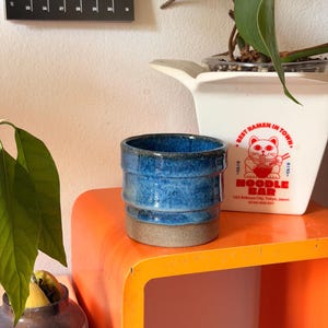 Peut inclure: Une tasse en céramique cylindrique présentant une section supérieure émaillée bleu vif avec une finition texturée et marbrée, contrastant avec une base grise mate non émaillée. La tasse a un design simple à parois droites.