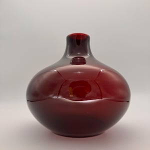 Puede incluir: Un jarrón de vidrio de color rojo intenso con un cuerpo redondeado y bulboso y un cuello estrecho y corto. El vidrio tiene un acabado liso y brillante que refleja la luz, creando reflejos luminosos en la superficie curva del recipiente.